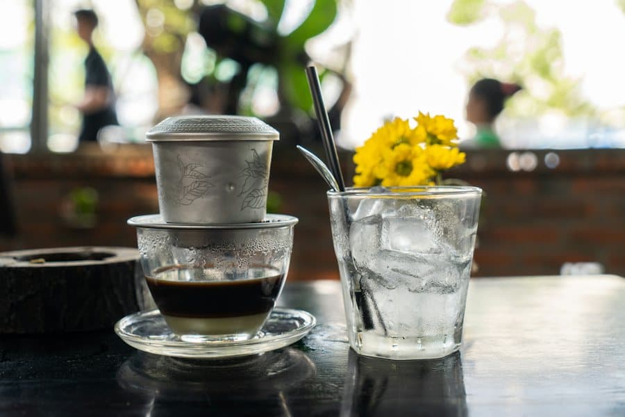 Ly cà phê phin đang nhỏ giọt, bên cạnh ly trà đá và bình hoa vàng