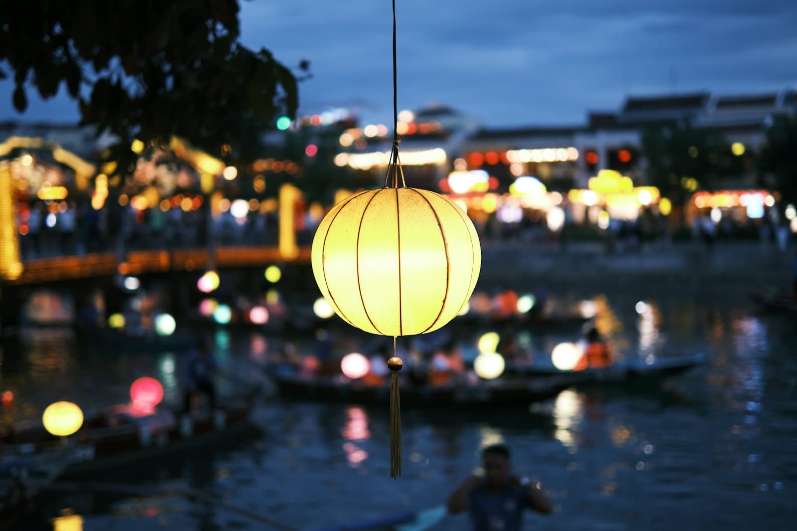 Một chiếc đèn lồng sáng treo bên bờ sông Hội An lúc chạng vạng, thuyền và đèn phố phía xa